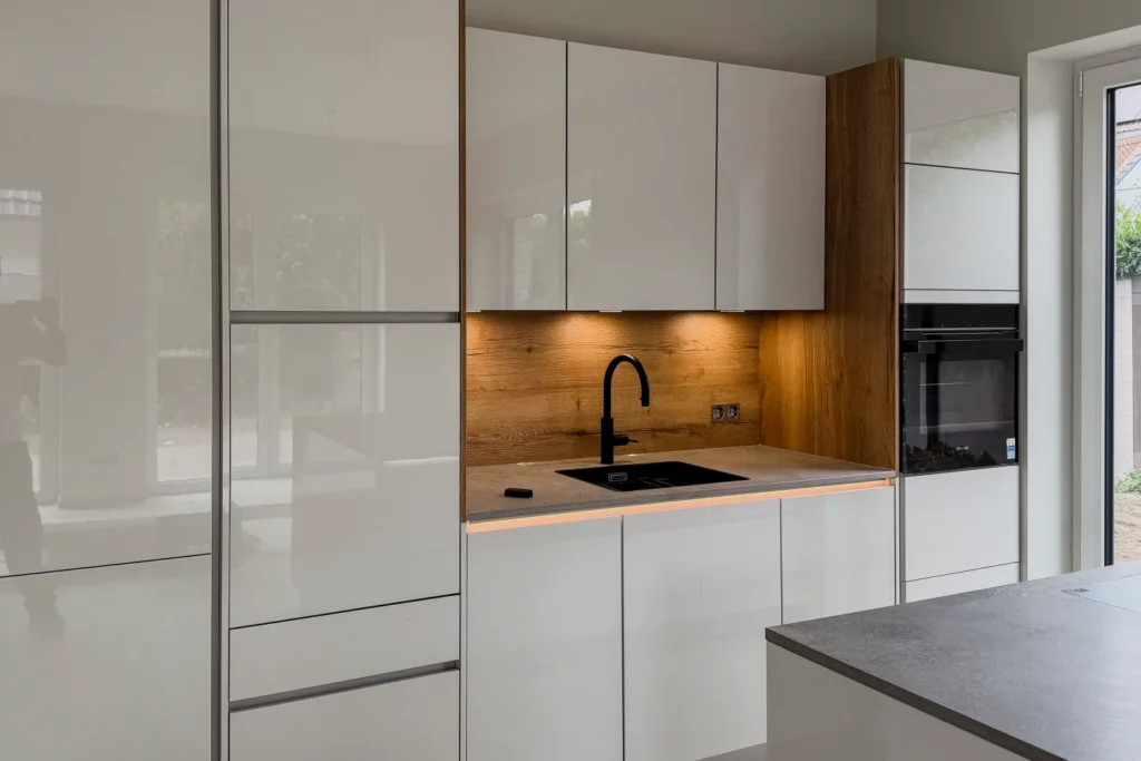 Moderne Küchenzeile mit weißen Fronten, Holzrückwand, schwarzem Kochfeld, Spülbecken und Einbau-Backofen.