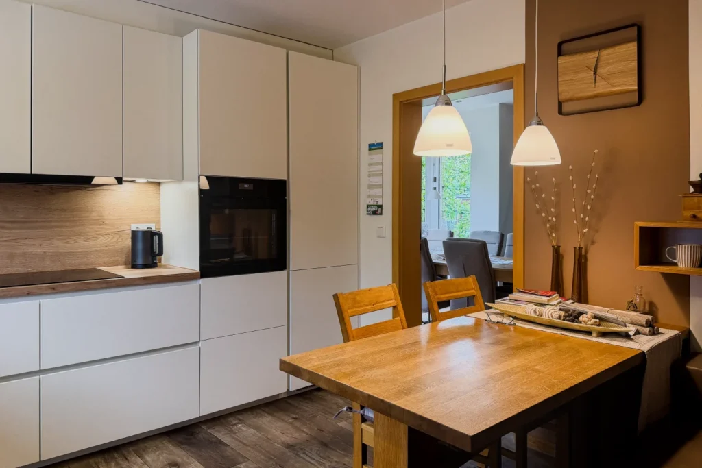 Helle Küchenzeile mit Hochschränken, Backofen, Holztisch und hängenden Lampen im Wohnküchenbereich.