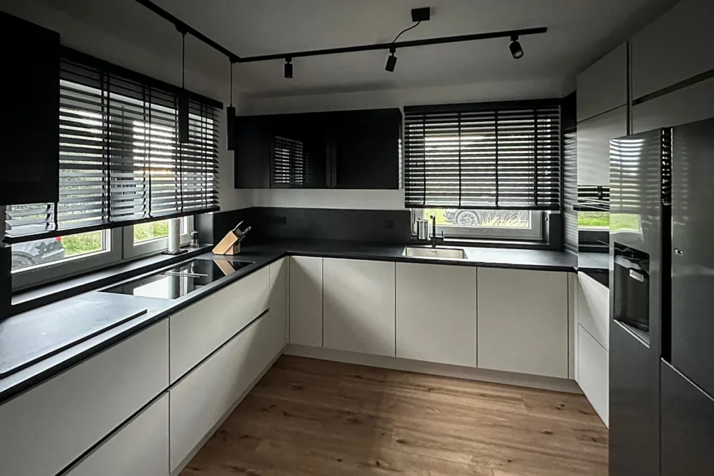Moderne L-förmige Einbauküche mit Spüle, Elektrogeräten, dunkler Decke und Jalousien über den Fenstern.