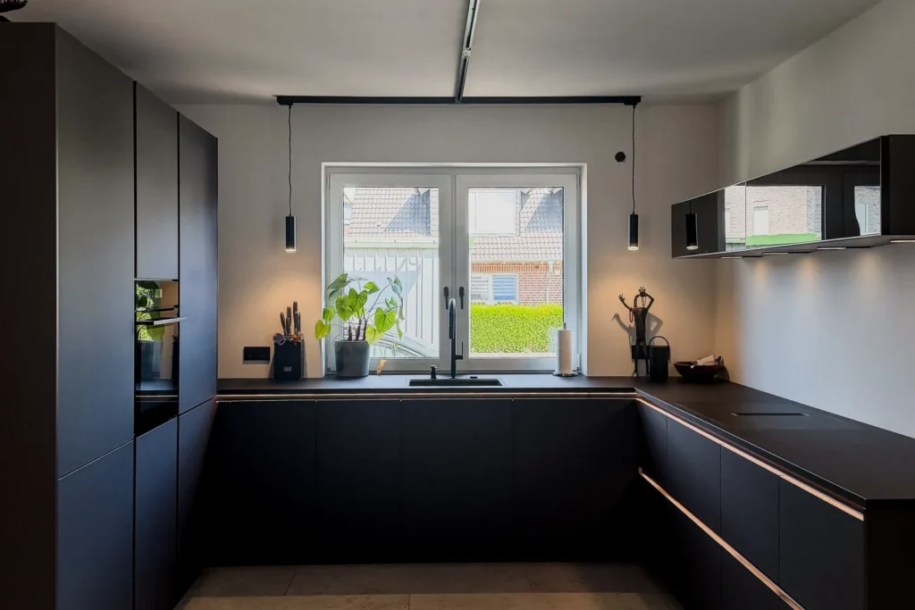 Dunkle Einbauküche mit Arbeitsplatte, Spüle am Fenster, seitlichen Schränken und Pflanzen im Innenraum.