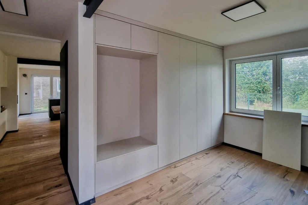 Einbauküche mit Hochschrank und Sideboard neben großen Fenstern, helles Zimmer mit Holzfußboden.