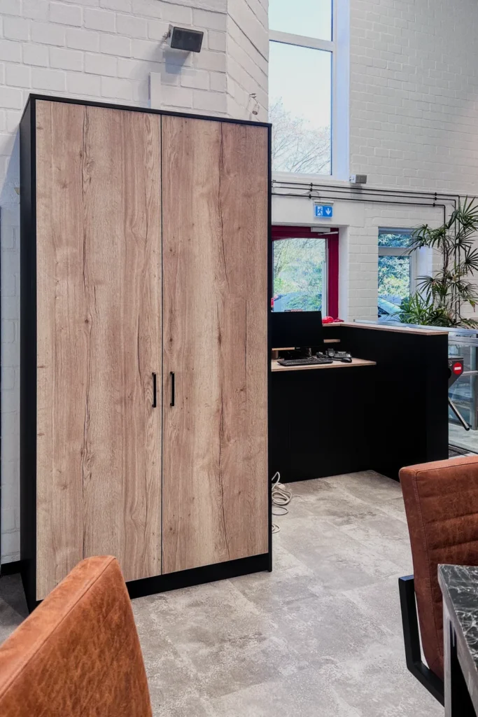 Holzschrank mit schwarzen Kanten und zwei Drehtüren, davor gepolsterter Stuhl, dahinter Küchenzeile mit Fenster.