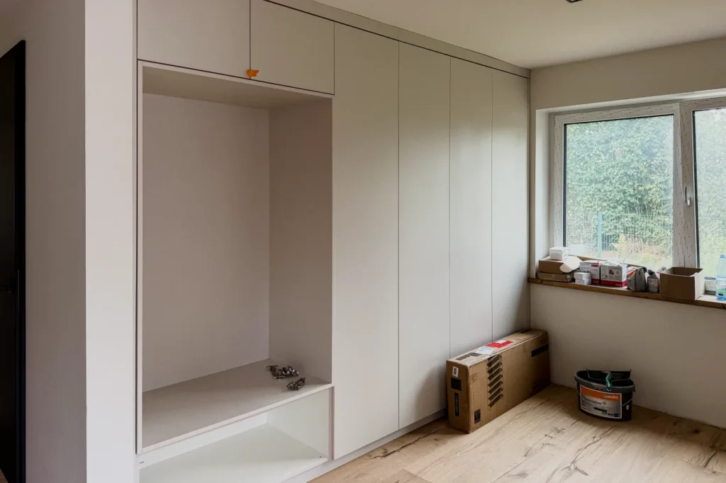 Weiße Einbauküche mit Schrankfronten und offener Nische neben Fenster, Boden mit Kisten und Farbeimer.