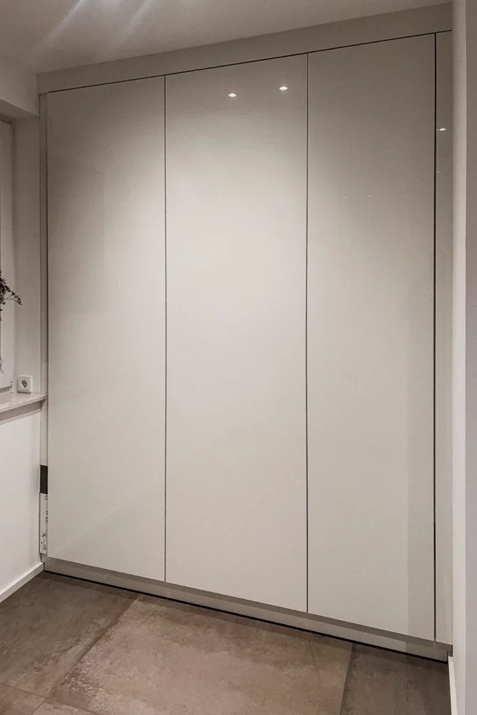 Einbauschrank mit drei glatten Schwebetüren ohne sichtbare Griffe in heller Wand, Boden mit Fliesen.