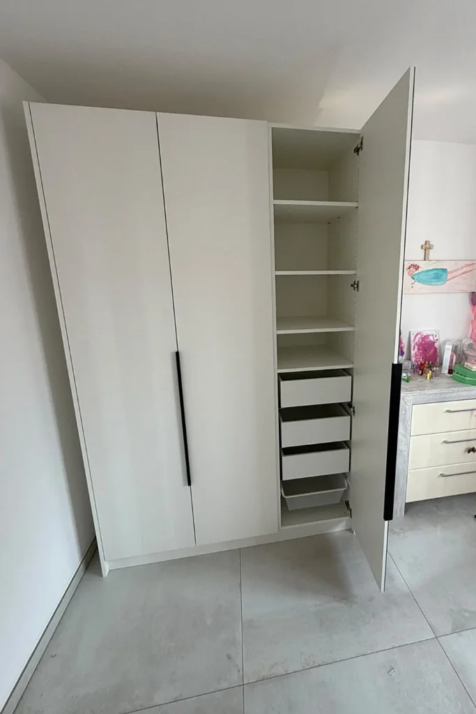 Offener weißer Küchenschrank mit Innenregalen und mehreren Schubladen, daneben eine Küchenzeile mit Arbeitsfläche.