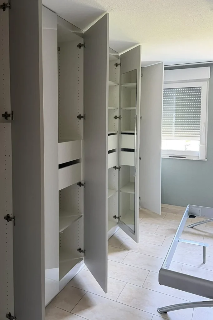 Offenes Küchenschrank-Fach mit Regalböden, Flurtürzeile und Fenster mit Rollos im Hintergrund.