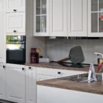 Küchenzeile in Magnolia mit Oberschränken, Backofen, Spüle und Arbeitsplatte mit Küchenutensilien.